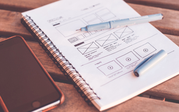 A notebook with wireframe sketches for web design, a pen, and a smartphone on a wooden table.