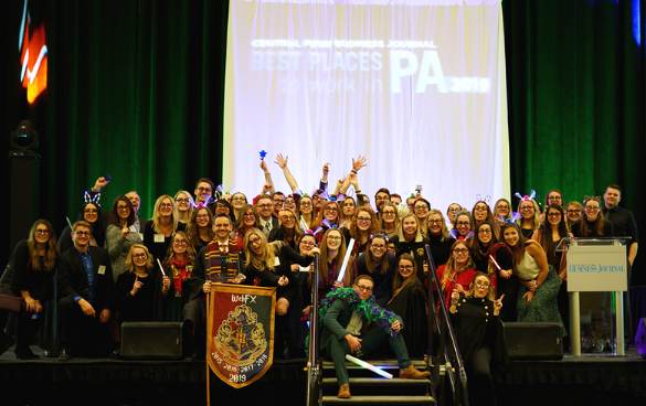 A cheerful group of people posing on a stage at an event celebrating 'BEST PLACES TO WORK IN PA 2019', with a banner and festive props.