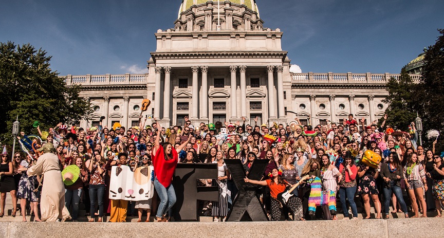 A diverse group of people in colorful costumes and holding props gathered on the steps of a grand capitol building, posing for a group photo in a festive atmosphere.