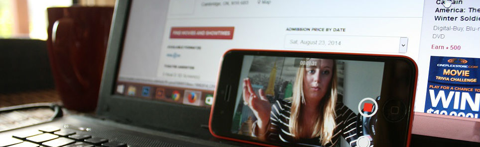 A laptop displaying a webpage and a red mug on a table, with a smartphone in front showing a woman on its screen as if on a video call.