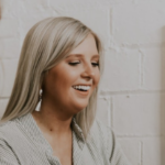 Two blonde women smiling and laughing together, one in a white top slightly out of focus and the other in a striped shirt in focus, in front of a plain wall and wooden cabinet.