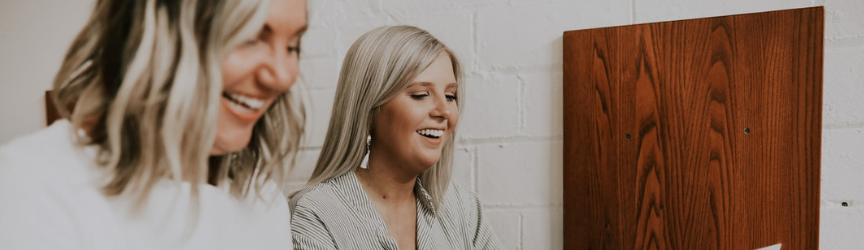 Two blonde women smiling and laughing together, one in a white top slightly out of focus and the other in a striped shirt in focus, in front of a plain wall and wooden cabinet.