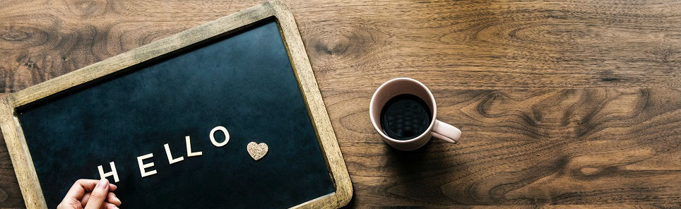 A chalkboard with the word 'HELLO' and a heart shape on it, a hand holding the board, and a cup of coffee on a wooden surface.