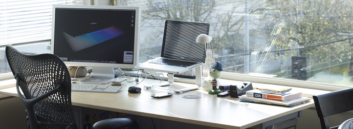 A tidy office desk with a dual monitor setup, including a large screen and a laptop on a stand, various office supplies, and a black chair, in a room with natural light from a large window.