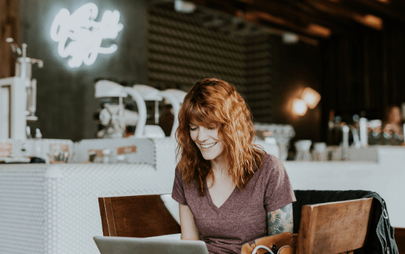 A smiling woman with reddish hair using a laptop at a wooden table in a rustic cafe, with a coffee machine and neon sign in the background.