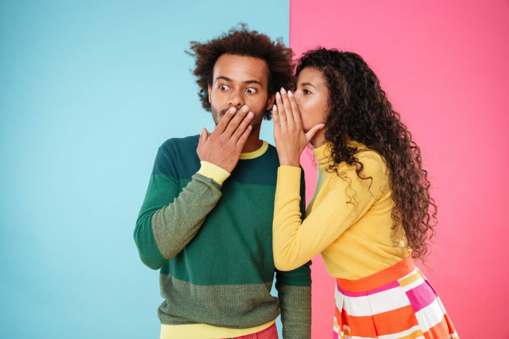 A woman whispering into a surprised man's ear against a half blue, half pink background.