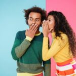A woman whispering into a surprised man's ear against a half blue, half pink background.