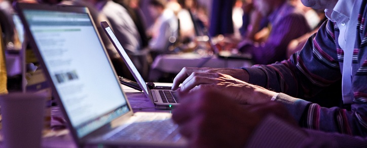 Close-up of a person's hands typing on a laptop with a screen displaying text, in a conference or workshop setting with other attendees in the background.