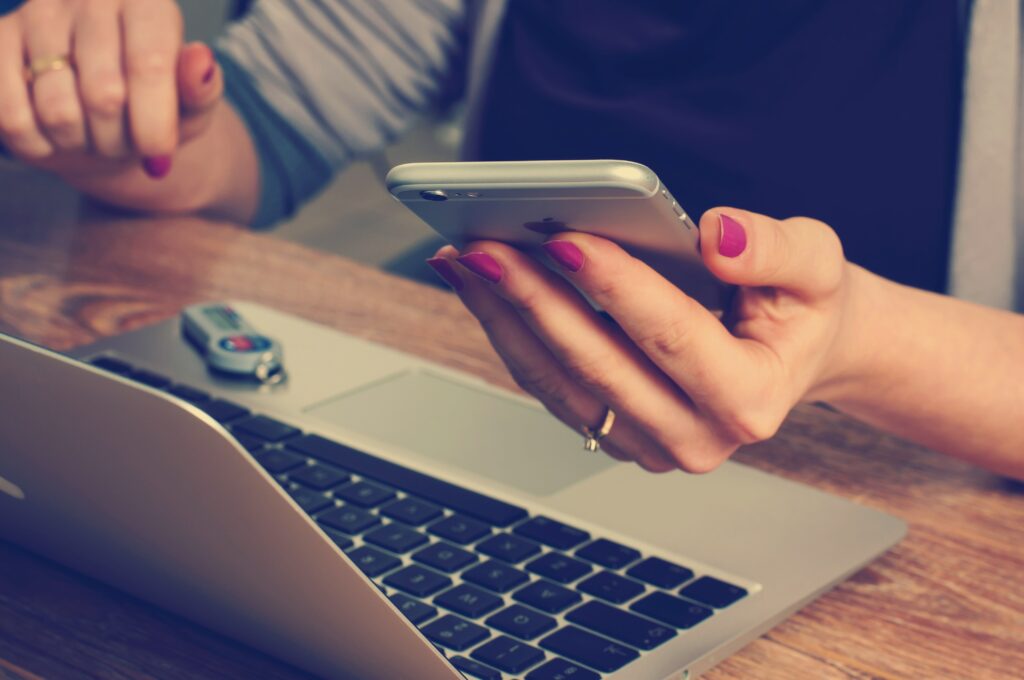 A person with purple nail polish using a smartphone with their laptop open and a car key fob on the desk.