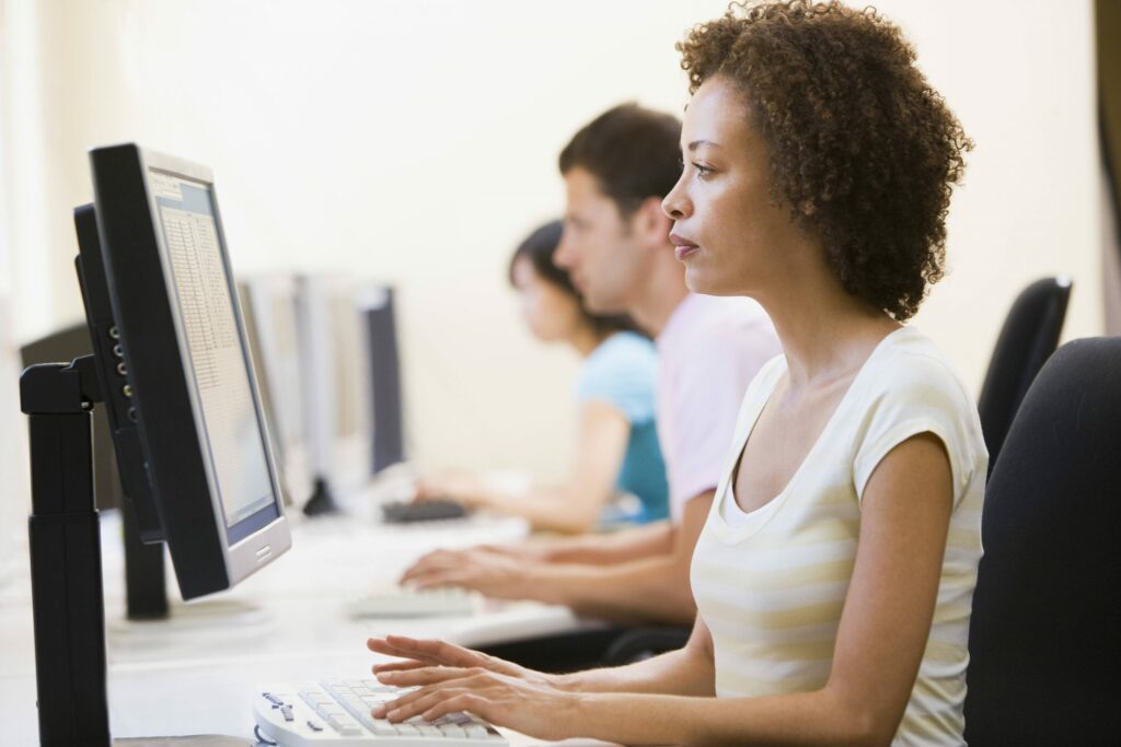 Three people using desktop computers in a row, possibly in a computer lab or office setting, with the woman in the foreground focused on her screen.