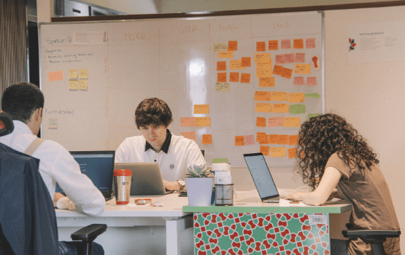 Three professionals working at a desk with laptops in a collaborative office setting, with a whiteboard covered in sticky notes in the background.