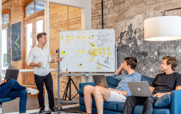 Three men in a casual office setting with one presenting at a whiteboard and the other two seated with laptops, engaged in a discussion.