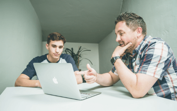 Two people, a young man and an adult man, are sitting at a table with a laptop. The adult is gesturing and appears to be explaining something to the younger man who is focused on the laptop screen.