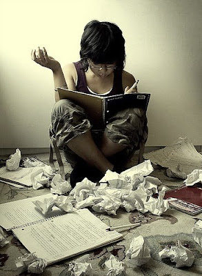 Person sitting on the floor, surrounded by crumpled paper, holding a pen and reading a notebook, suggesting a study session or creative process.