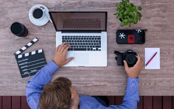 A man sits in front of a laptop with a clapboard to his left and camera to his right