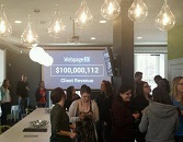 A group of people networking in a room with a presentation screen showing 'WidgetPro RX' and '$100,000,113 Global Revenue'. Modern lighting fixtures are visible overhead.