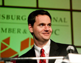 Smiling man in a suit speaking at a podium with a microphone, with a 'PITTSBURGH REGIONAL CHAMBER OF COMMERCE' banner in the background.