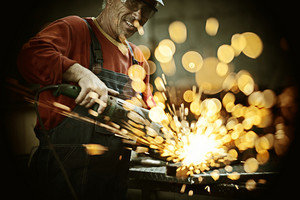 A smiling worker in safety goggles and overalls uses a grinding tool on metal, creating a vibrant shower of sparks.