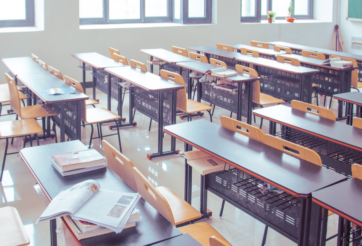 An empty classroom with rows of paired desks and attached benches, scattered books and papers on the desks, and natural light from side windows.