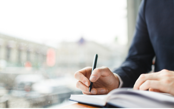 Close-up of a person in a dark blue suit writing in a notebook, with a blurred interior background suggesting a well-lit space.