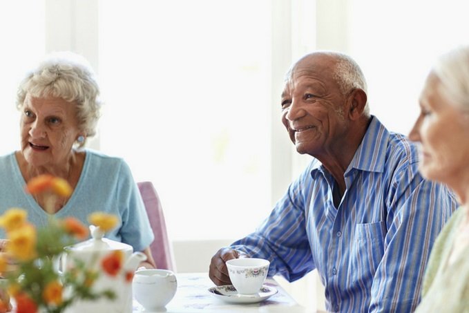 Three elderly individuals enjoying a tea time together, with a smiling man in a blue striped shirt holding a teacup, an engaged woman in a light blue sweater, and another partially visible woman, in a bright room with flowers on the table.