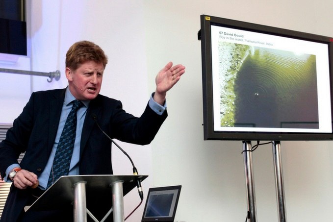 A man in a suit presenting at a podium, gesturing towards a screen displaying a photograph related to the Yamuna River in India.