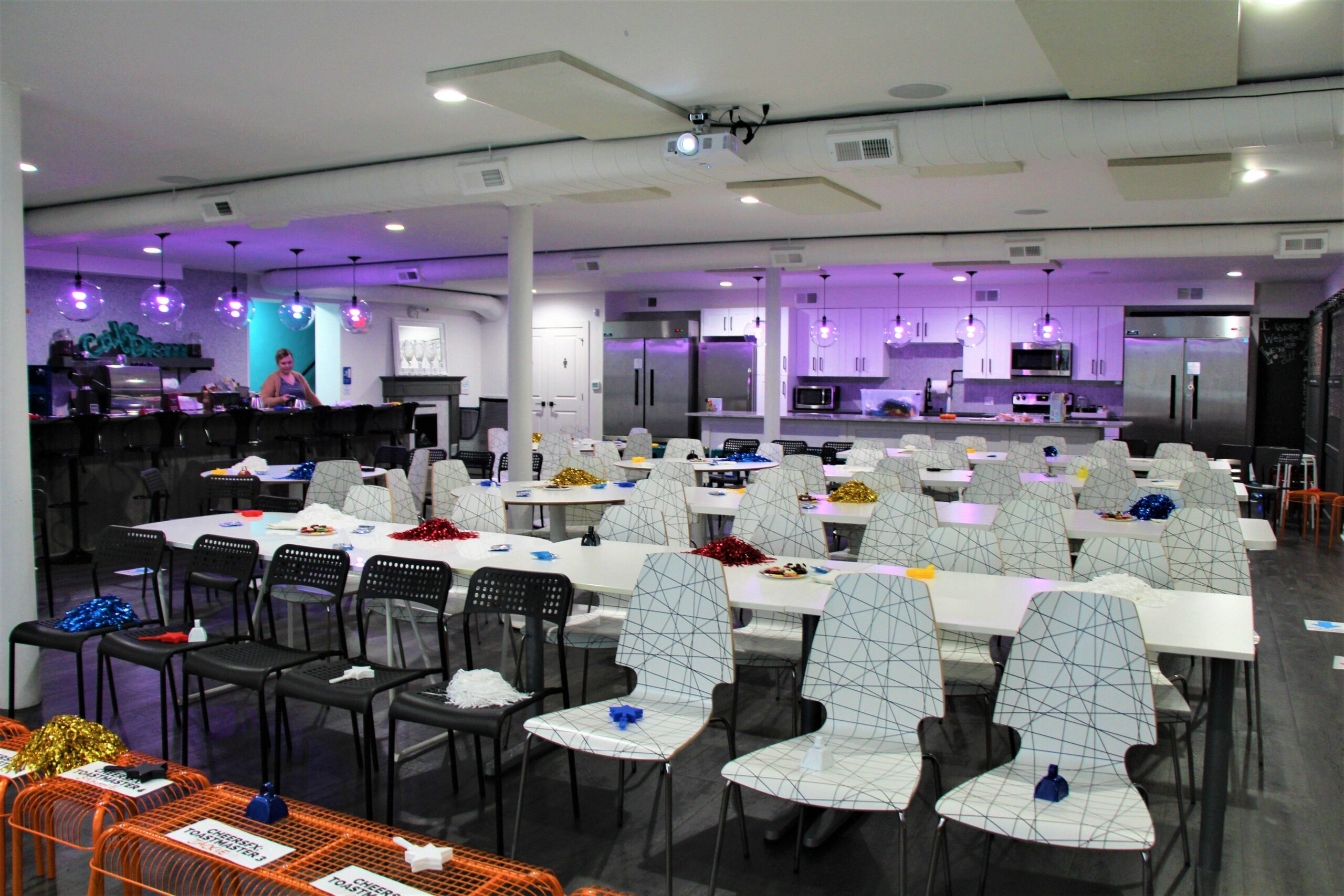 A modern event space with tables set up for a party, decorated with colorful pom-poms and party hats, and a kitchen area in the background with one person standing behind the bar counter.