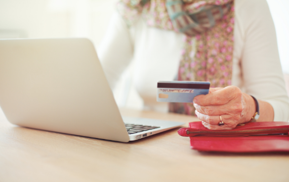 An older adult with a scarf and a white long-sleeve shirt is holding a credit card while using a laptop, with a red wallet on the table.