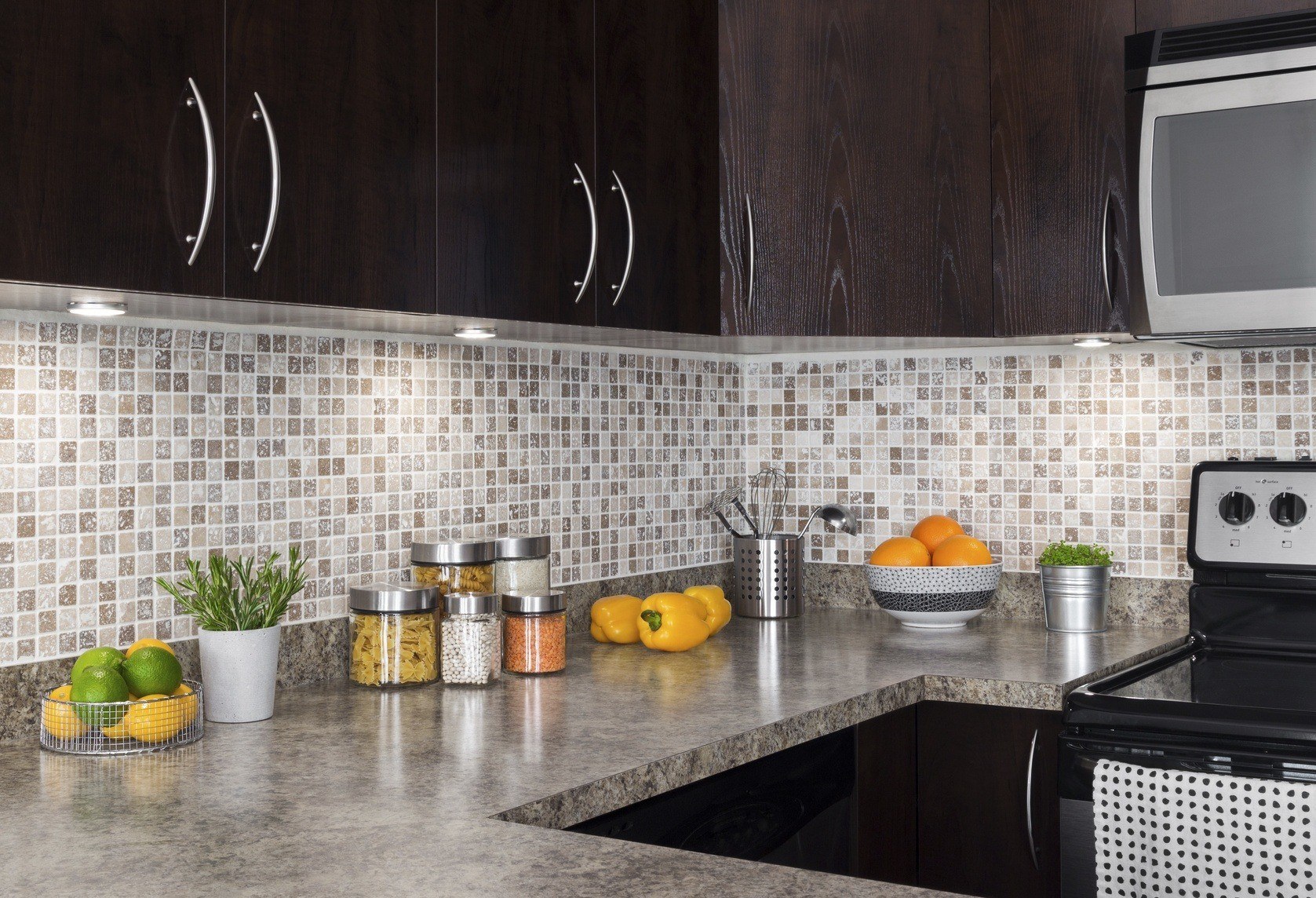 Modern kitchen with dark wood cabinets, mosaic tile backsplash, and granite countertop featuring a microwave, stove, and various fruits, herbs, and cooking utensils.