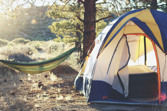 A camping setup with a dome tent and an air mattress in a forested area, next to a green hammock tied between two trees, bathed in warm sunlight.