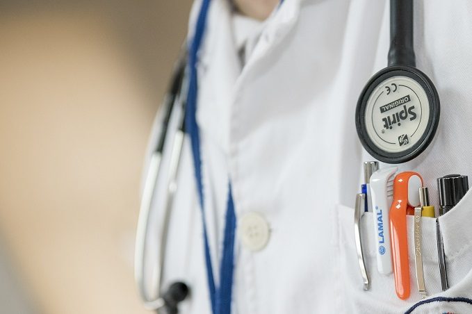 Close-up of a medical professional wearing a white lab coat with a 'Spirit' stethoscope around the neck and various tools such as pens, a penlight, and scissors in the coat pocket.