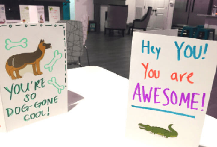 Two greeting cards on a table; one with a dog illustration saying 'YOU'RE SO DOG GONE COOL!' and another with the message 'Hey YOU! You are AWESOME!' with a crocodile illustration, in a blurred office setting.