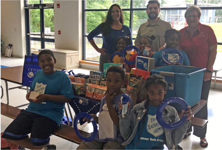 Three adults and four children posing with a shopping cart full of food donations inside a building, with some wearing shirts with 'The Salvation Army' logo, indicating a charitable event.