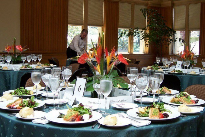 A banquet hall with round tables set for a formal meal, complete with floral centerpieces, place settings, and a waiter in the background.