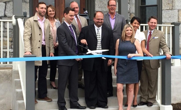 A group of eight people at a ribbon-cutting ceremony in front of a building, with two men in the center holding large scissors ready to cut a blue ribbon.