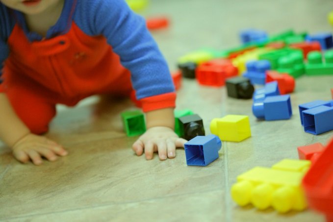 A young child in a red overall is playing with colorful building blocks on the floor, reaching for a blue block.