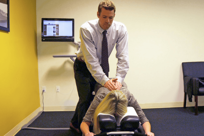 A chiropractor in a shirt and tie performing a back adjustment on a female patient lying face down on a chiropractic table in a clinic.