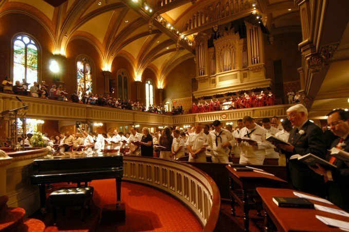 Choir in white robes standing in a church with attendees seated in pews and on a balcony during a service, with arched ceilings and stained glass windows, and a grand piano in the foreground.