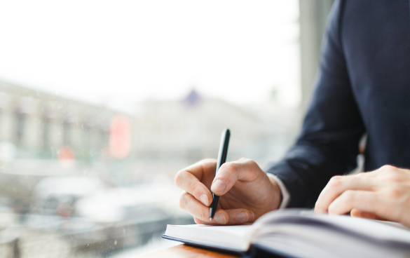 Close-up of a person in a dark blue suit writing in a notebook with a black pen, with an out-of-focus cityscape visible through a window in the background.