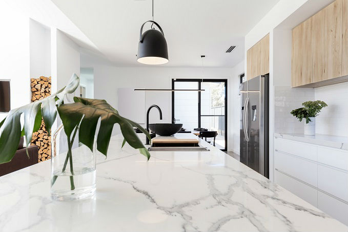 Modern kitchen with white marble countertops, white cabinetry, and stainless steel appliances, featuring a monstera leaf in a vase, a black pendant light, and an open-plan living area in the background.