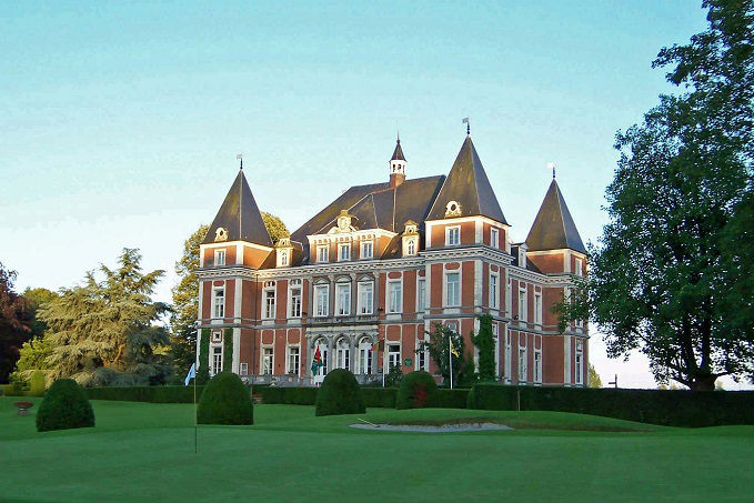 A large chateau-style building with red brick facade and pointed roofs, surrounded by a manicured lawn under a clear blue sky.