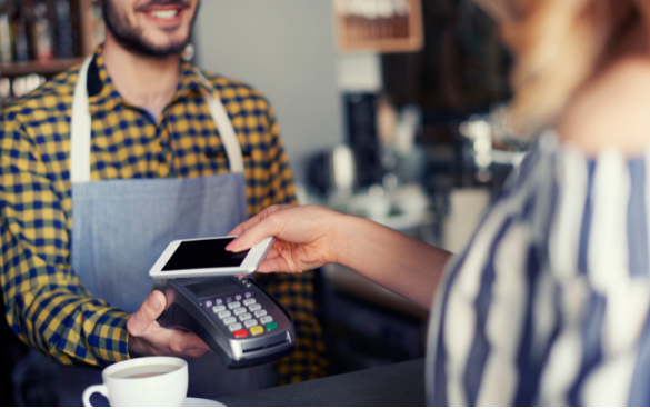 A smiling server in a checkered apron holds a card payment terminal while a customer uses a smartphone for contactless payment at a café counter, with a coffee cup visible in the foreground.