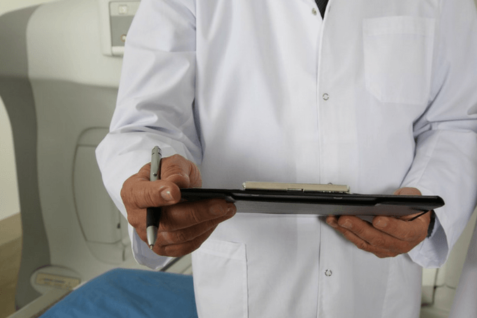 Healthcare professional in a white lab coat holding a clipboard and pen in front of medical imaging equipment.