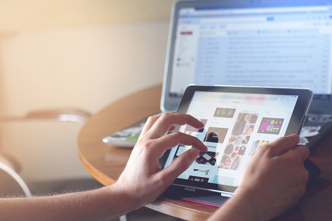 Person using a tablet with a webpage open while an open laptop is in the background on a wooden desk.