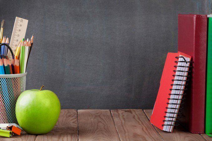 A green apple, colored pencils in a mesh container, a ruler, scissors, and a red spiral notebook leaning against two large books on a wooden table with a chalkboard background.