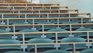 An empty lecture hall with rows of green upholstered seats and wooden desks, and a green chalkboard in the background.