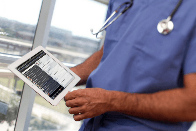 Healthcare professional in blue scrubs with a stethoscope holding a tablet displaying a medical application, standing beside a window overlooking a cityscape.