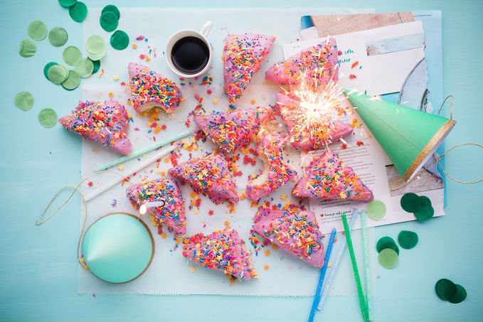 A celebratory arrangement with pink frosted pastries covered in rainbow sprinkles and a lit sparkler, surrounded by a party hat, a cup of coffee, confetti, and papers on a light blue surface.