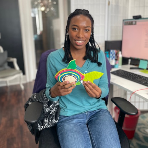 Smiling woman sitting in an office chair, holding a colorful paper fish craft, with a desk and computer monitor in the background.