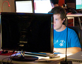 Person in a blue t-shirt concentrating on a computer screen in a dimly lit room with ambient lighting indicative of a gaming event or hackathon.